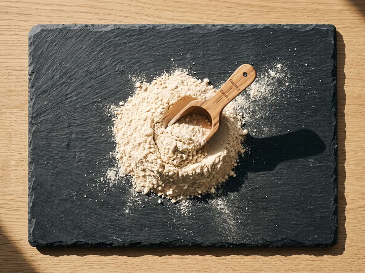 A heap of pale cream digestive enzyme powder on dark slate, a wooden scoop half-buried in it